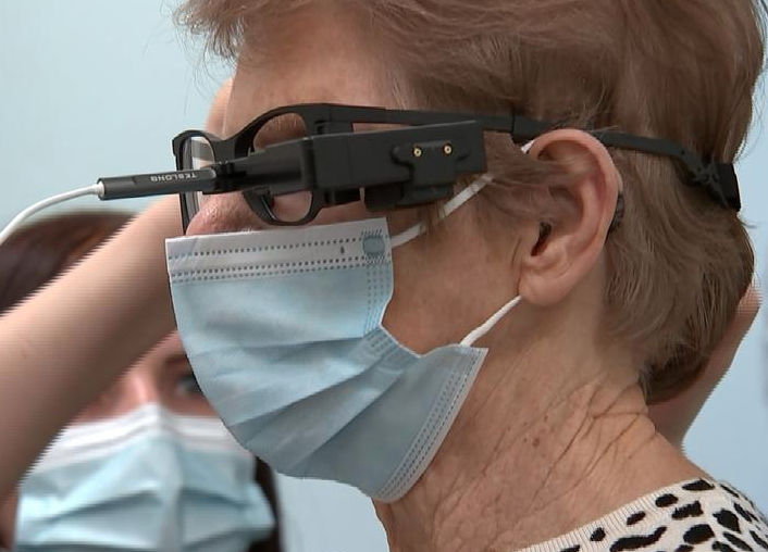 Image of lady in the foreground wearing a face mask, having a bionic eye fitted by a specialist sitting beside her