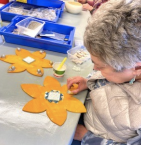 Image of EYECAN member Carol sitting at a table working on a sunflower template made of wood and painted yellow. She is sticking small mosaics on to the template. 