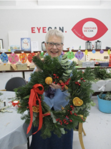Image of EYECAN member Enid standing up and proudly holding her Christmas Wreath. The green wreath is decorated with red ribbon and red berries with slices of dried orange. 