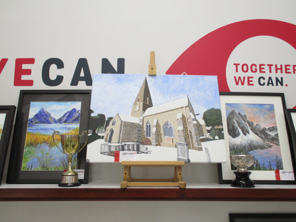 Image shows 3 landscape paintings and two trophies in front of them. The painting on the left is of a lake with reeds in it and mountains in the background; The middle painting is of St Clement's Church; And the painting on the far right is of a lake surrounded by mountains, snow and trees