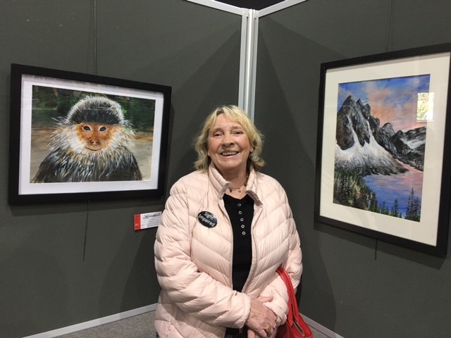 Image shows Laura standing between two paintings, one is of a monkey and the other of a lake surrounded by mountains, snow and trees