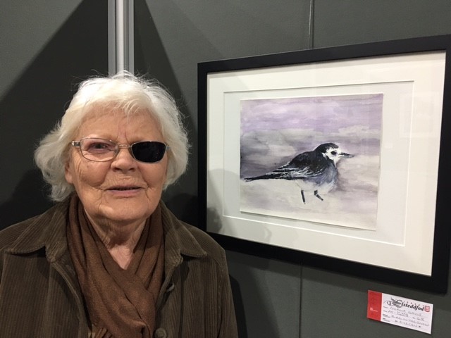 Image shows Norma, who is wearing an eyepatch, posing next to a painting of a small black and white bird