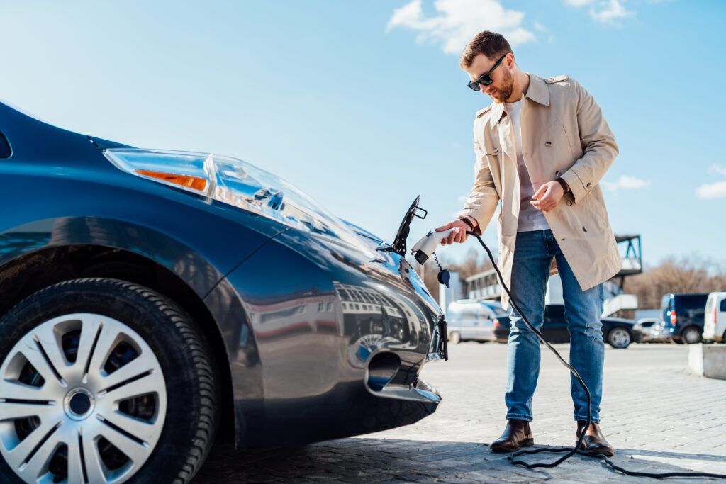 gentleman wearing beige jacket and blue jeans holding an electric charging plug. He is standing by a black car