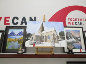  Image shows 3 landscape paintings and two trophies in front of them. The painting on the left is of a lake with reeds in it and mountains in the background; The middle painting is of St Clement’s Church; And the painting on the far right is of a lake surrounded by mountains, snow and trees.