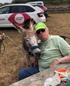 Image shows EYECAN member Ed sitting on a chair next to a brown and white donkey. The donkey’s head is leaning on Ed’s shoulder. An EYECAN car is in the background