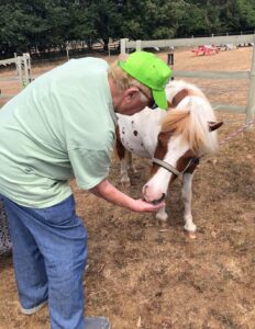 Image shows EYECAN member Ed dressed in jeans and a green T-shirt. He is bending down to feed a brown and white pony. The pony is taking food from Ed’s hand