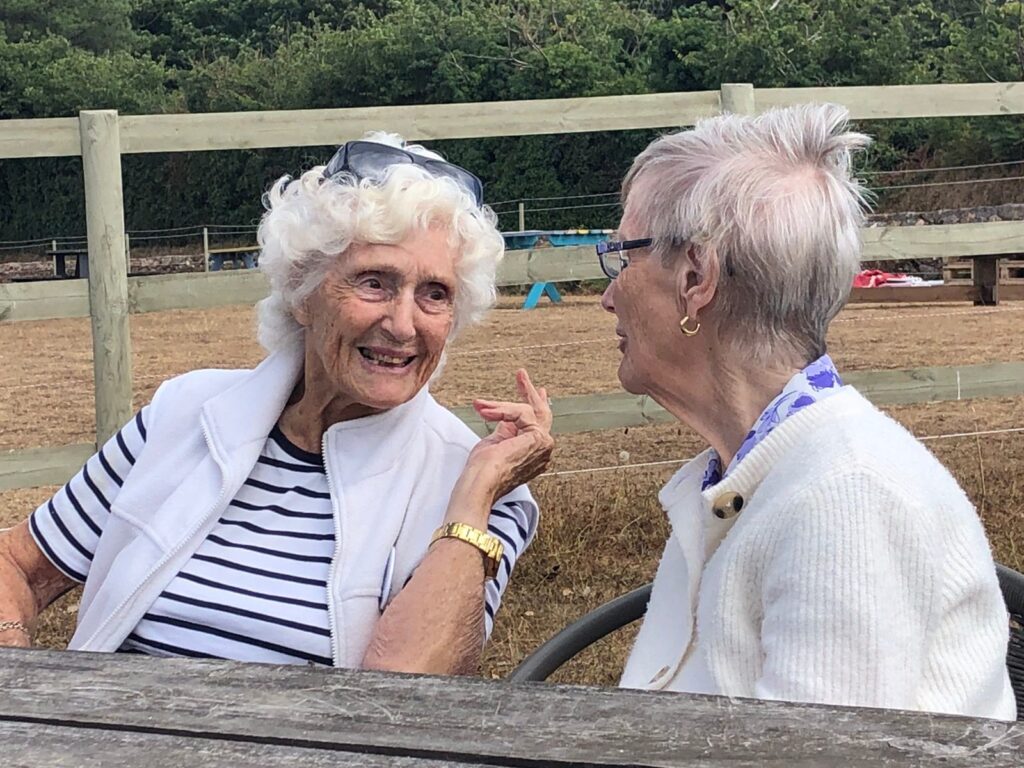 Image shows two lady members of EYECAN engaged in conversation. One is wearing a white cardigan and the other lady is wearing a blue and white striped top with a pair of sunglasses on her head