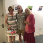 Image shows Jane, Graham and Shanon standing together at the art exhibition in the jersey library. Jane is wearing a multicoloured dress, Graham is wearing a checkered shirt and dark brown shorts, Shanon is wearing pink clothes. 