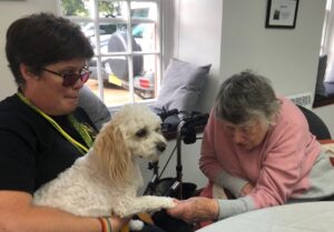 Image shows Simone who is wearing a pink and grey jumper. She is leaning forward in her chair and holding Basil’s paw in her left hand. Sarah is wearing black and is sitting down next to Simone with Basil on her lap. Basil is a small white therapy dog.
