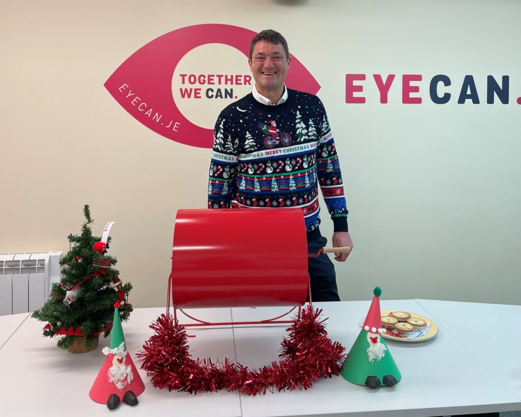Image shows EYECAN President Ed Daubeney wearing a Christmas jumper depicting Santa Claus riding a bike in a night scene. He is standing behind the red raffle ticket tombola