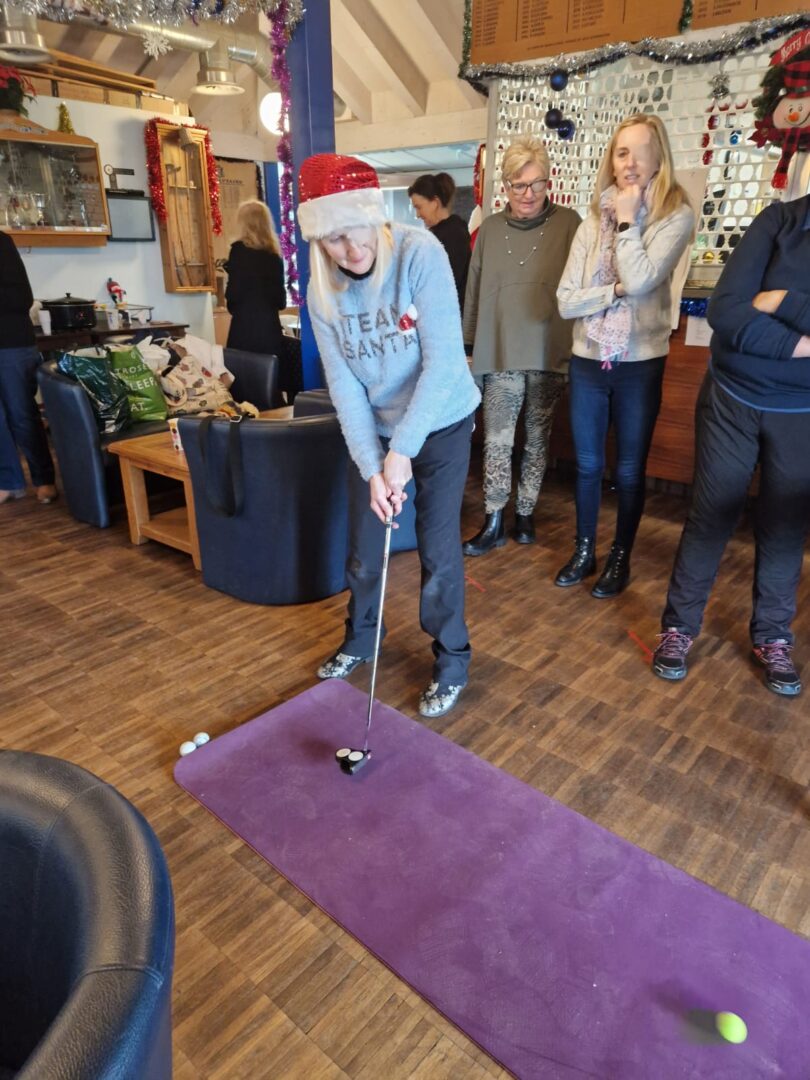 Image shows Mary's friend wearing a red and white Christmas hat and Christmas sweater, she is putting a golf ball while wearing an eye patch