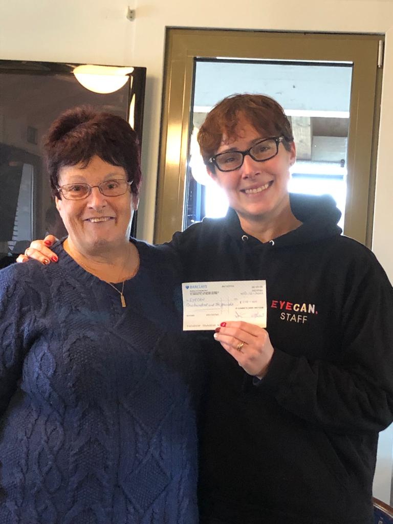 Image shows EYECAN's Sight Support Officer Agnetta with her arm around Mary. Mary is wearing a blue sweater and smiling at the camera. Agnetta is in her EYECAN uniform and is holding a cheque towards the camera.