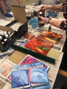 Image shows a table covered with newspaper, on the table is a black box with different coloured paint tubes and two paintings. One painting has red, yellow and green colours and the other a mixture of yellow, orange, red and black colours.