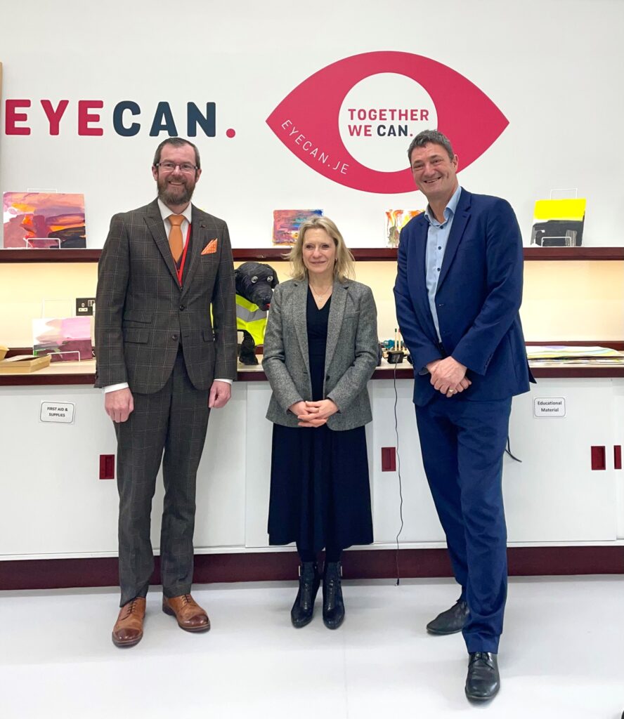 Image shows Deputy Elaine Millar wearing a black dress and grey jacket standing between Deputy Malcolm Ferey wearing a brown suit and orange tie and Ed Daubeney who is dressed in a blue suit. They are standing in front of the EYECAN logo. 