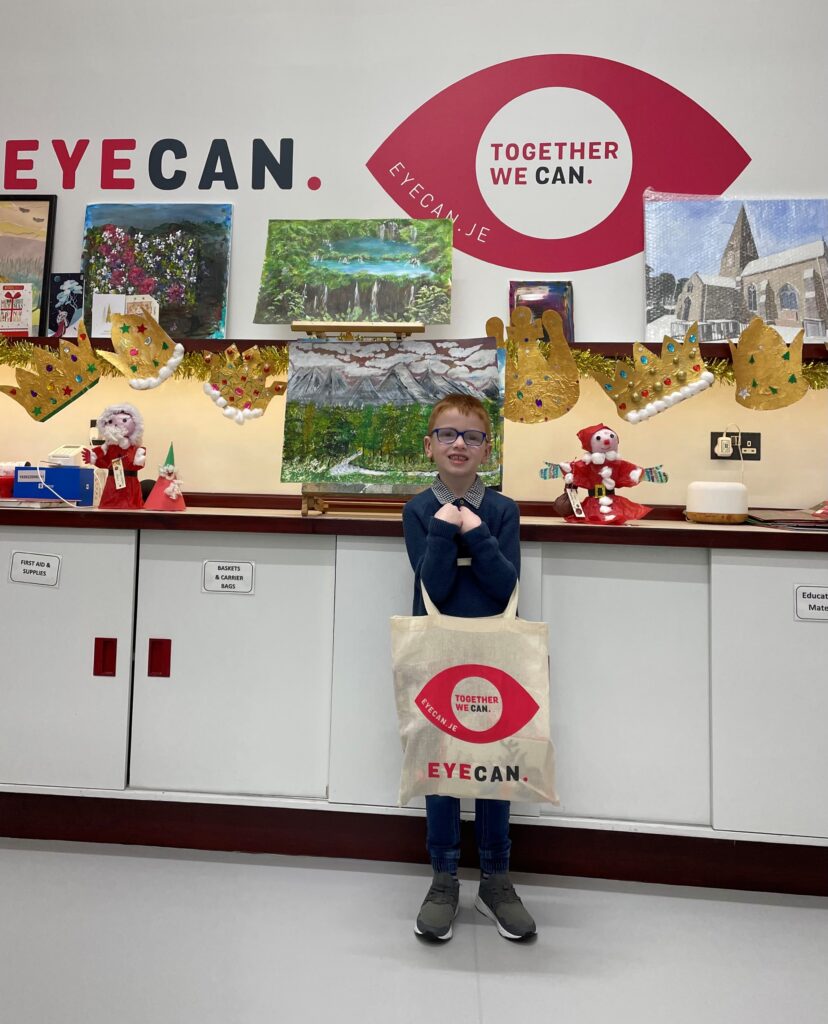 Image of James standing and holding his EYECAN goodie bag. He is dressed in a blue jumper and trousers and he is wearing blue glasses. There are paintings and Christmas decorations behind him