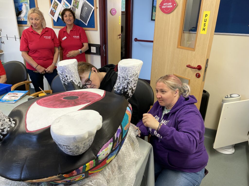 Image shows our art club members getting the tortoise ready, there are two EYECAN volunteers standing behind the tortoise and two art club members painting the bottom of the tortoise sculpture. The tortoise has a big red and white eye painted on the bottom of it's shell.