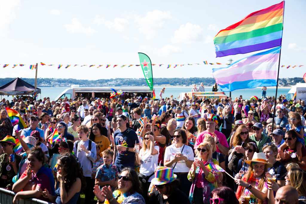Image of a crowd celebrating the Pride festival in Jersey with the sea in the background. The following wording in white is written across the image: "Supporting Islanders with Sight Impairment at Channel Islands Pride 2023, Saturday 16th September.