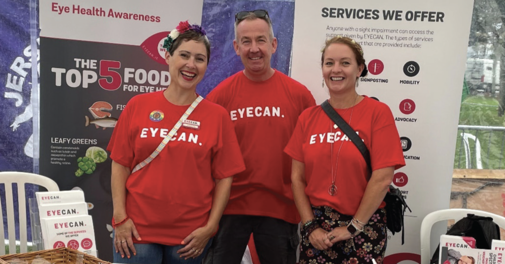 Image shows Mark Coxshall standing in between Agnetta and Emmaline. They are standing in front of EYECAN pop up banners and wearing red EYECAN T-Shirts. They were attending CI Pride event 2023