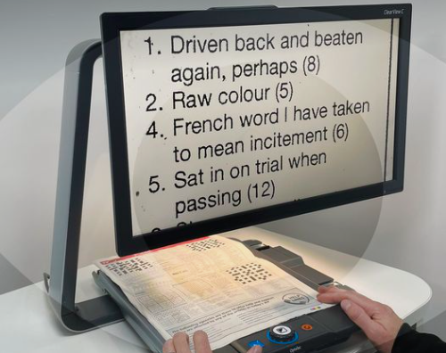 Image shows video magnifier placed on a white table. The screen is displaying an image of words that are questions from a crossword puzzle. The words have been magnified